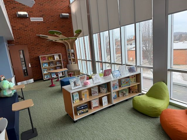 Children's reading area with picture books in book cases, soft seating, a tree book shelf in the corner, and small tables